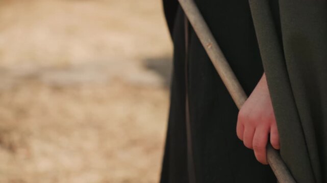 Tight outdoor view of lower torso and hand holding wooden staff while heavy dark green cloak drapes with deep vertical folds, creating rustic texture, grounded posture, and quiet sense of resolve