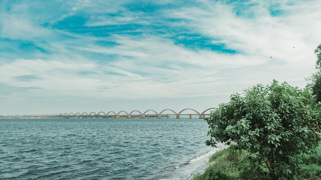 Aerial view of the Godavari Arch Bridge spanning the wide Godavari River with lush green trees in the foreground under a bright blue sky in Rajamahendravaram, Andhra Pradesh, India.
