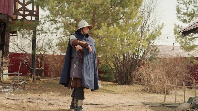Historical-style performer stands in weathered outdoor courtyard holding vertical polearm, cloak resting over armor while gentle breeze adds slight movement, with rural textures and natural daylight
