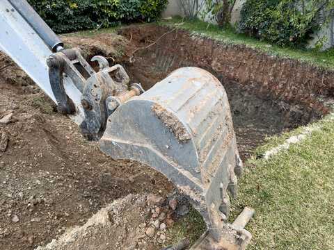 Mud covered excavator bucket digging a deep foundation pit in a residential garden