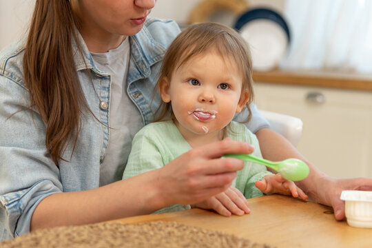 Happy family at home. Mother feeding her baby girl from spoon in kitchen. Little toddler child with messy funny face eats healthy food at home. Young woman mom giving food to kid daughter