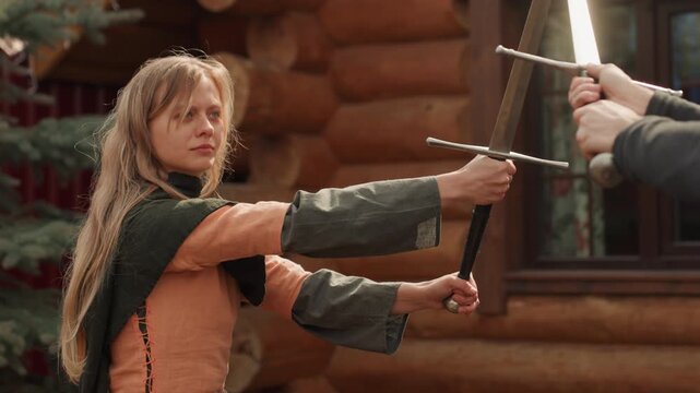 Young woman in braided blonde hair and green cloak braces with spear, parrying incoming strike while bearded man lifts weapon for upward swing beside rustic log cabin in warm daylight