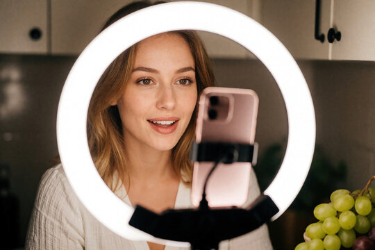 Young caucasian woman makeup live stream using smartphone, ring light. Beautiful girl vlogger records video blog social media content at home. Female influencer talks to followers online through