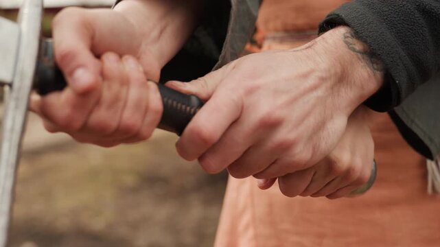 Close-up of hands gripping and adjusting leather wrapped sword hilt, with twisting motion, thumb pressure, and forearm tension near crossguard and pommel in outdoor historical reenactment setting
