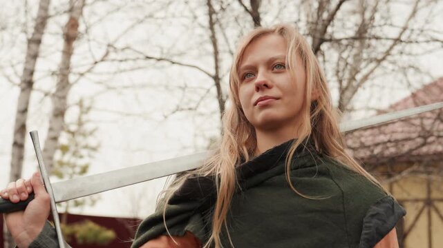 Young woman stands in residential yard holding polearm near shoulder, chin raised and steady gaze showing confidence, with layered costume, soft breeze, birch tree, fence, muted sky