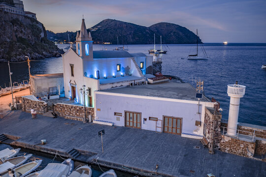 Aerial view of the illuminated Church of the Souls in Purgatory situated by the harbor with moored boats and calm sea at dusk in Lipari, Sicily, Italy.