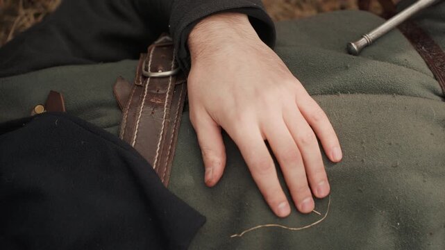 Close-up view of adult hand gently resting on olive canvas backpack strap, fingers relaxed while making subtle adjustment, with visible leather trim, stitching, metal hardware, worn texture, and