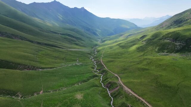 Stunning drone shot of a lush alpine valley with a stream, rural path, and massive ridges in summer