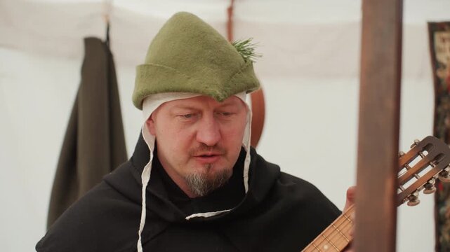 Middle aged man in green felt hat and dark cloak holds wooden stringed instrument at chest height while adjusting strings and looking down with concentrated calm expression inside softly lit tented