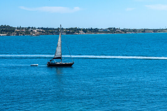 a view of the sky and the blue sea of Syracuse, criss-crossed by the wake of a boat and a sailing boat 