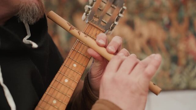 Tight horizontal close-up shows musician playing small wooden fretted instrument with metal slide, left hand pressing neck while right hand glides over strings, captured in warm outdoor light with