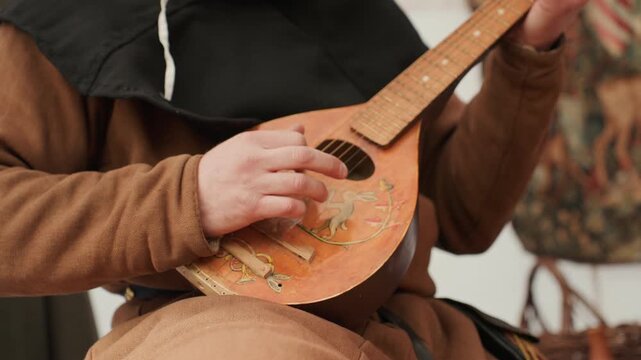 Hands actively strum and finger carved round-bodied wooden string instrument with floral decoration, captured under warm diffused indoor light with visible chord changes, rhythmic movement, and