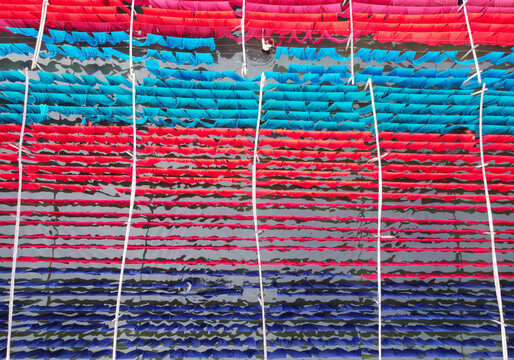 Aerial view of vibrant red, blue, and turquoise fabrics hanging on bamboo poles to dry over water, with a worker tending to the textiles in Narayanganj, Dhaka, Bangladesh.