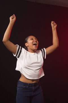 African American woman raising arms, shouting in studio wearing white cropped tee lit by red rim