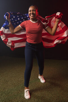 African American woman holding and spreading United States flag in studio on turf, wearing red tee