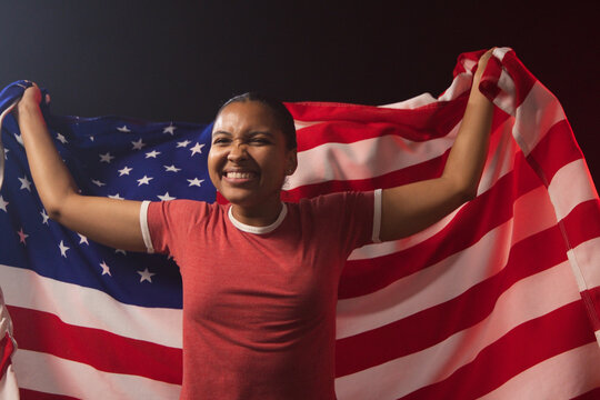 African American woman holding US flag behind shoulders, wearing red ringer tee and studs, smiling