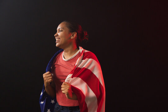 African American woman standing, holding US flag over red T-shirt and dark jeans in studio, smiling