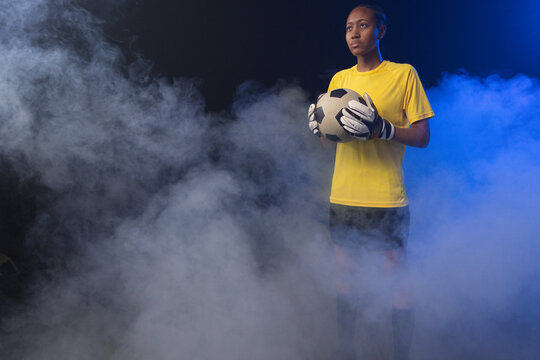 Female goalkeeper standing amid fog on stage holding soccer ball, yellow jersey, gloves, copy space