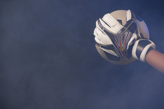 Gloved hand gripping soccer ball against deep blue haze, showing wrist strap, copy space