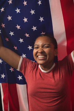 African American woman standing before American flag holding flag, smiling in red ringer tee, studs