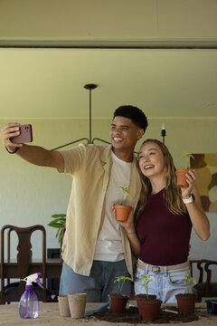 Couple posing and taking selfie at dining table, holding seedlings with purple smartphone