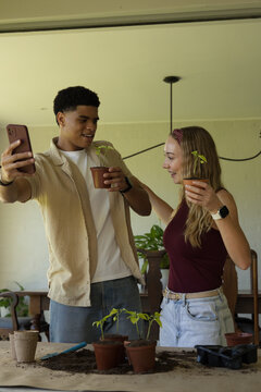 Couple taking selfie while potting seedlings at wooden table with smartphone and trowel