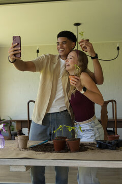 Diverse couple smiling, taking selfie while potting seedlings on wood table with purple smartphone