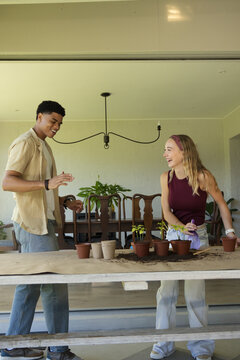 Couple potting seedlings and laughing on covered patio at long wooden table with terracotta pots