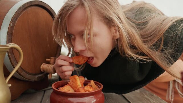 Woman leans over rustic wooden table, reaches for pastry in clay bowl, then brings it toward mouth with focused expression, while barrel and brass pitcher frame warm natural light and homestyle