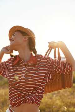 Young woman in a striped shirt and straw hat basks in sunset, carrying a wicker basket, enjoying the warm breeze. Outdoor lifestyle moment for fashion, travel, or leisure.