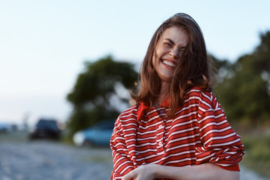 A smiling woman in a red striped shirt stands outdoors by the coast, capturing a casual lifestyle moment. Bright daylight highlights carefree style and warm mood.