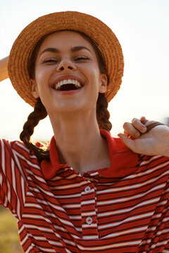 Young woman wearing a straw hat laughs with joy outdoors, in a red striped shirt, braids visible, sunlit scene conveying carefree summer energy and candid happiness.