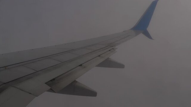 Airplane wing navigating through thick grey fog and dense clouds in overcast sky, viewed from cabin window in real time with soft daylight.