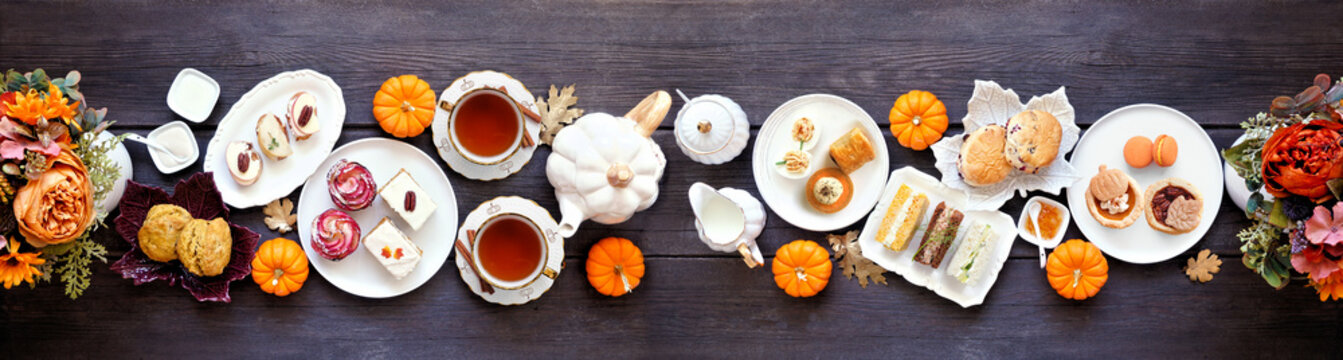 Autumn theme afternoon tea table scene against a dark wood banner background. Pumpkin and apple theme savory finger foods, sandwiches, scones, and desserts. Overhead view with copy space.