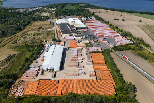 Aerial view of a large industrial brick storage facility with rows of orange construction materials, warehouses, and surrounding fields near a lake in Konin, Wielkopolskie, Poland.