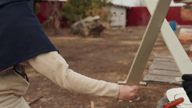 Children in cloaks spar with wooden swords in rustic backyard, one stepping back while other swings, creating lively outdoor motion, friendly interaction, and imaginative play in daylight