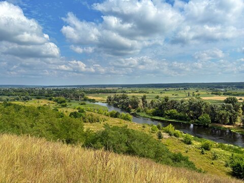 High-angle panoramic view of a calm winding river flowing through a lush green valley with golden grass on the hill under a cloudy blue sky.