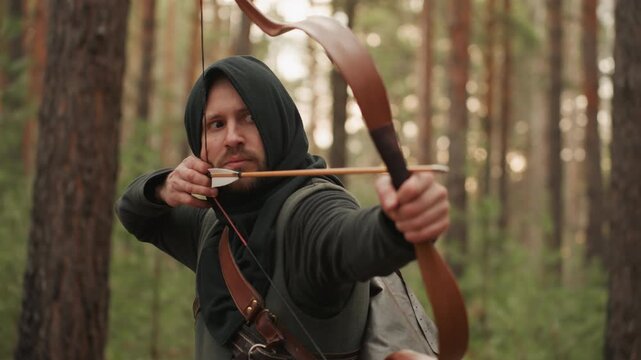 Hooded archer in medieval style clothing draws arrow to cheek, then lowers bow while standing in pine forest with focused determined expression, leather gear, soft light, tall trees