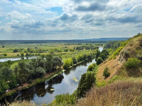 High-angle view of a calm river winding through dense green trees and summer fields under a soft cloudy sky.