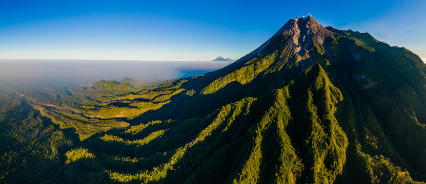 Aerial view of Merapi volcano with lush green slopes, rugged volcanic terrain, and clear blue sky in Yogyakarta, Indonesia.