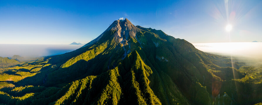 Aerial view of Merapi volcano with its rugged volcanic slopes, lush green forests, and a clear blue sky during a bright sunny day in Yogyakarta, Indonesia.
