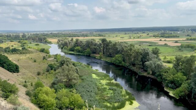 Panoramic stock video of a calm winding river flowing through a green countryside valley with lush trees and fields under a cloudy sky.
