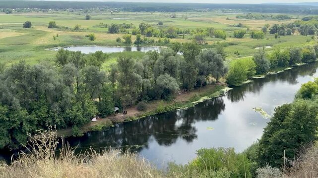 Panoramic stock video of a calm winding river flowing through a green countryside valley with lush trees and fields under a cloudy sky.
