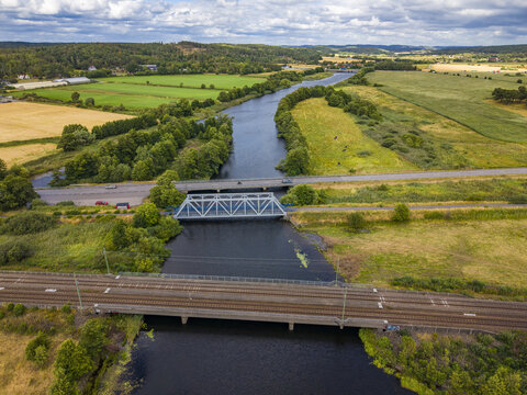 Aerial view of the Viskan river flowing through the rural landscape, featuring a railway bridge and a road bridge surrounded by green fields and trees in Askloster, Halland County, Sweden.
