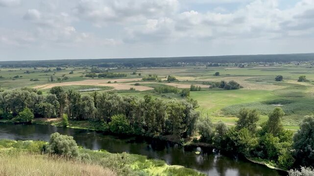 Panoramic stock video of a calm winding river flowing through a green countryside valley with lush trees and fields under a cloudy sky.
