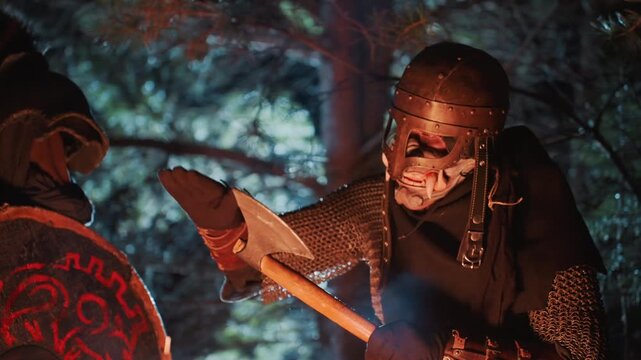 Armored knight in helmet and chainmail leans toward illuminated parchment while companion in cloak stands nearby, torchlight mixing with moonlight among trees for tense adventurous mood
