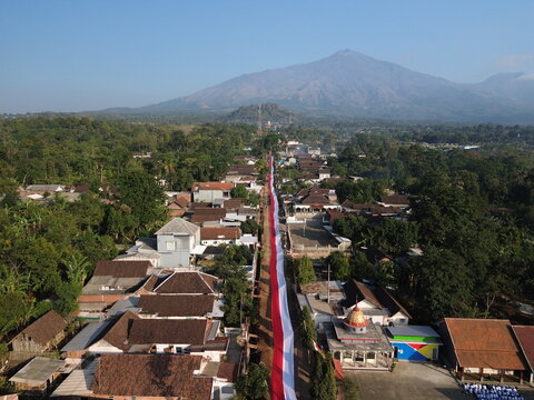 Aerial view of a long red and white Indonesian flag stretched along a village street with houses and lush greenery, with a mountain in the background, Wonosari, East Java, Indonesia.