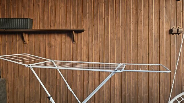 A white metal drying rack holds a crumpled grey garment and a small white cloth against warm wooden panels; a dark shelf and metal fittings accent the rustic setting