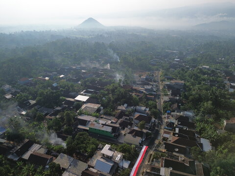 Aerial view of the town of Wonosari nestled among lush tropical trees and misty hills with residential rooftops and a red and white flag visible in Wonosari, East Java, Indonesia.