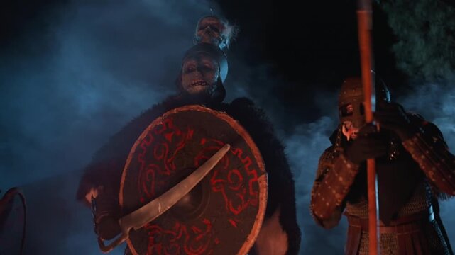 Two armored combatants press forward through dense smoke, shield bearer facing camera with ornate round shield while companion crouches with spear raised, creating tense dramatic motion
